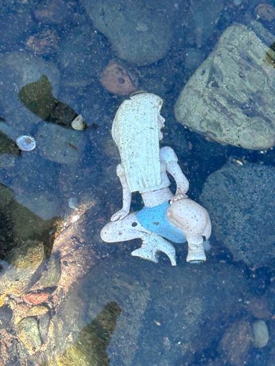 Photograph of a plastic stripper toy laying among some rocks underwater. The stripper’s head is turned at an impossible angle so that its long blonde hair falls down over its chest and its face can only be seen in profile, mouth open. It wears a white midriff-exposing shirt, blue hot pants, and white high heels. It squats over the heels with hands on its thighs.