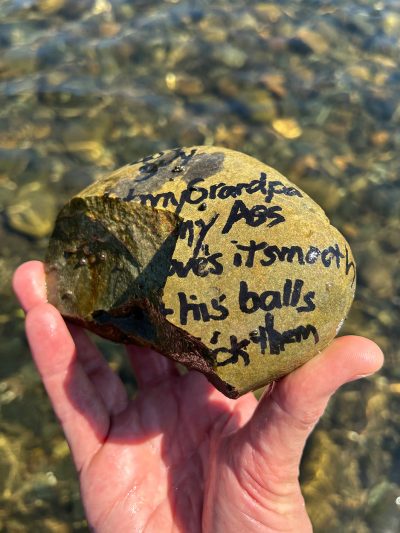 Photo of a hand holding a round river rock approximately five inches long and three inches tall. Someone wrote on the rock in permanent marker but the rock is broken, eliminating some of the text. The remainder reads, “[ … ] guy [ … ] my Grandpa [ … ]t my Ass [ … ]ove’s it smooth [ … ] his balls [ … ]ick them[.]”