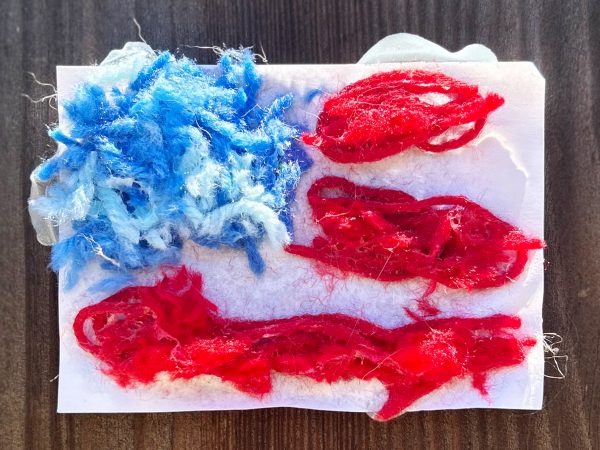 Photo of a small piece of white paper on which blue and red yard is glued in an approximation of the flag of the United States of America. There is a square of pinched-together blue yard in the top left corner, and three lines of red yarn with spaces in between them so that the white of the paper can be seen. This makeshift flag sits on top of a piece of dull-brown wood. “‘The truth, my friends, is that America is exceptional because here, nothing is fixed into place. The frontier may be closed, we may have walked on the moon, but the work of fulfilling the values first enshrined in the Declaration of Independence–that work endures, my friends, and it belongs to us all.’”--Zohran Mamdani.