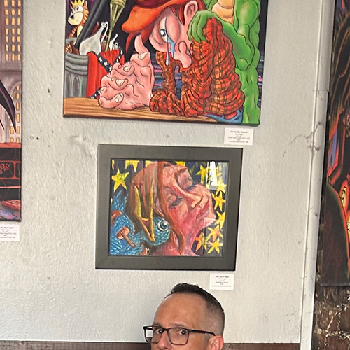 A photograph of Sacramento-based artist Jesse Baggs below various works of his art at Weatherstone during the August, 2026 You Are Here art show opening, although Baggs can only be seen from the nose up.