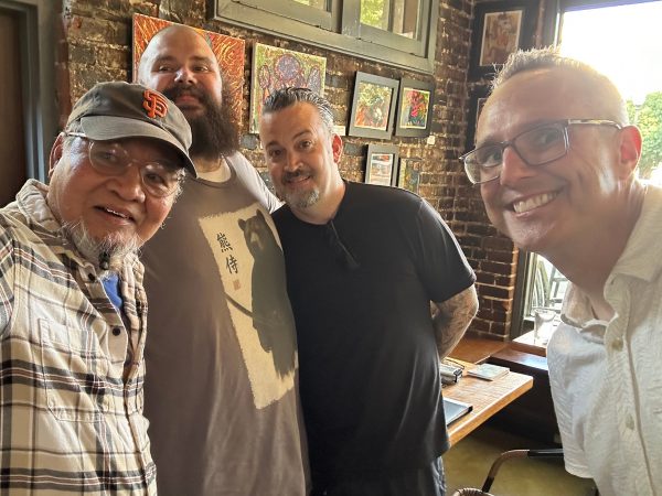 Photograph of Sacramento-based artists Raul Lacanilao, Gabriel Sanford, Emery Ensign, and Jesse Baggs standing in front of various works of art by Baggs for the August, 2026 You Are Here art show at Weatherstone. Both Lacanilao and Baggs are attempting to take a selfie at the same time, so Lacanilao and Sanford look at Lacanilao’s camera, while Ensign and Baggs look at Baggs’s.