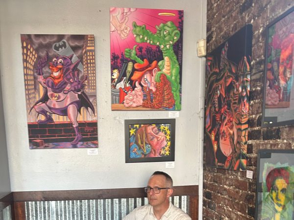 A photograph of Sacramento-based artist Jesse Baggs sitting below various works of his art at Weatherstone during the August, 2026 You Are Here art show opening.