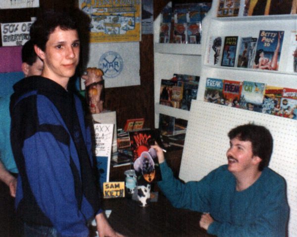 Photograph of Sacramento-based artist and cartoonist Jesse Baggs at age 14 in Sacramento comic-book store World’s Best Comics. Baggs stands at a table wearing a purple-and-black hoodie and looks to his right at someone out of frame. On the other side of the table, comic-book artist Sam Keith smiles at Baggs while holding up a copy of “The Maxx” #1 that he just signed. In the background is a large Barney Rubble toy and various comic books, including issues of “Superman,” “The Fabulous Furry Freak Brothers,” “Motor City Comics,” and “Betty Page in 3-D.”