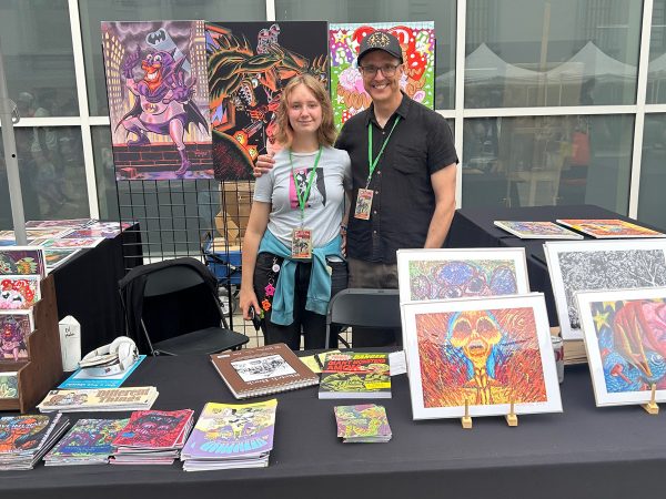 Photograph of Sacramento-based artist and cartoonist Jesse Baggs and his daughter at their table for CrockerCon 2026 at the Crocker Art Museum. Various prints, comics, stickers, and original art cover the table and hang from a rack behind them. Baggs’s daughter wears a Marceline / Princess Bubblegum shirt and handmade candy jewelry. Baggs wears a black button-up shirt and a black baseball cap featuring two dancing skeletons in gold and text that reads, “Let’s get weird.”