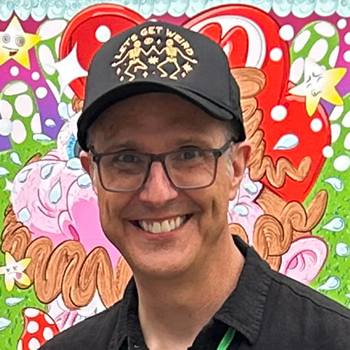 Photograph of Sacramento-based artist and cartoonist Jesse Baggs standing in front of his digital painting “Goomin’ Up that Hill [A Deal with Toad],” which depicts a sweaty Super Mario who has eaten too many mushrooms. Baggs wears a black button-up shirt and a black baseball cap featuring two dancing skeletons in gold and text that reads, “Let’s get weird.”